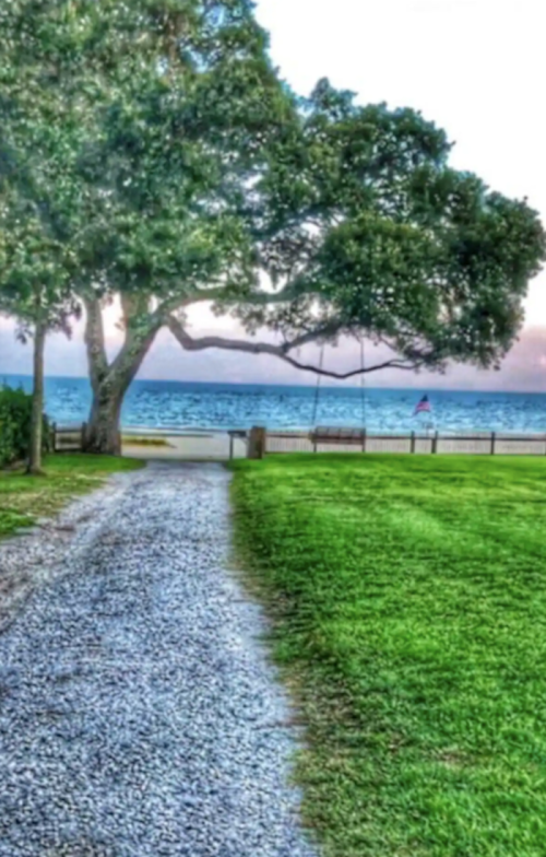 A gravel path leads to a serene waterfront view, with a swing under a large tree and an American flag in the distance.