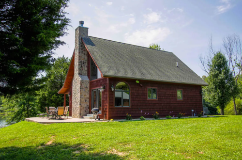 A charming wooden cabin with a stone chimney, surrounded by greenery and near a tranquil body of water.