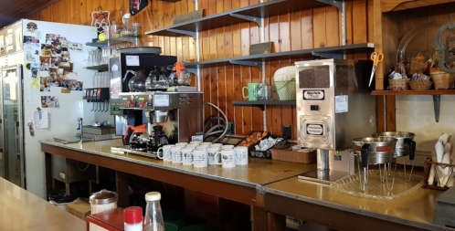 A cozy café interior featuring a coffee machine, shelves, mugs, and a display of baked goods.