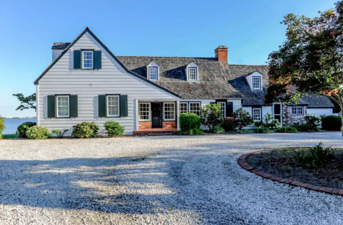 A charming gray house with a shingled roof, surrounded by greenery and a gravel driveway, near a body of water.