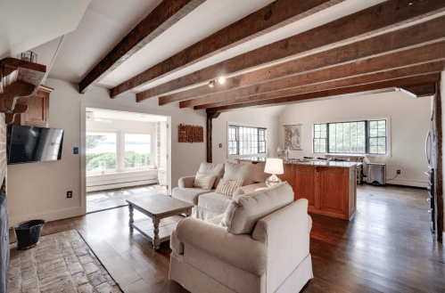 Cozy living room with wooden beams, light-colored furniture, and an open layout leading to a kitchen and scenic view.