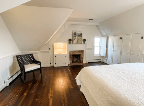 Cozy bedroom with a white quilt, wooden floors, a fireplace, and a chair near a slanted ceiling.