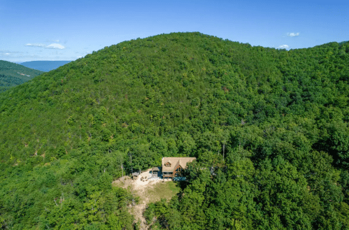 Aerial view of a cabin nestled in a lush green forested hillside under a clear blue sky.