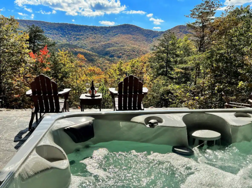 A hot tub with a scenic mountain view, surrounded by trees and two chairs with a table, under a blue sky.