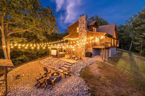 A cozy cabin at dusk, adorned with string lights, featuring a fire pit and surrounded by trees.