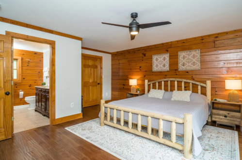 Cozy bedroom with wooden walls, a bed, nightstands, and a door leading to a bathroom. Natural light and warm decor.
