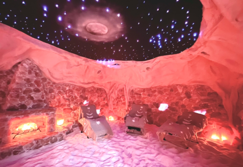 A cozy salt room with stone walls, two lounge chairs, and a starry ceiling, illuminated by soft lights and candles.