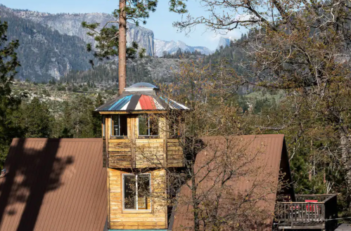 A wooden tower with a colorful roof stands among trees, overlooking mountains and a clear sky.