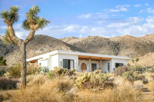 A modern white house surrounded by desert vegetation and mountains under a blue sky with scattered clouds.