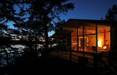 A cozy cabin by the water at dusk, illuminated from within, surrounded by trees and a serene lake view.