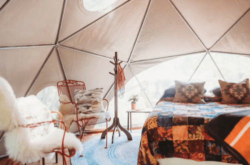 Cozy interior of a geodesic dome tent featuring a bed, chairs, and decorative elements. Natural light filters in.
