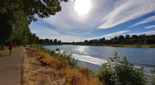 A scenic riverside path with a jogger, surrounded by trees and a bright sky reflecting on the water.