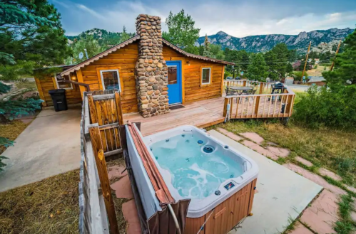 A cozy wooden cabin with a hot tub on a deck, surrounded by trees and mountains in the background.