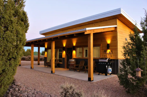 A modern wooden cabin with a porch, surrounded by gravel and shrubs, illuminated in the evening light.