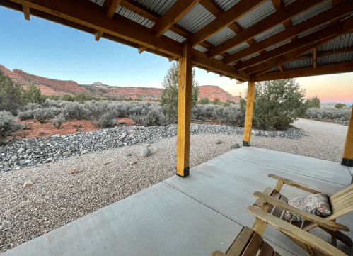 View from a porch featuring a gravel path, shrubs, and distant red rock formations under a pastel sky.