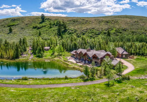 A scenic view of a large house by a pond, surrounded by lush greenery and rolling hills under a blue sky.