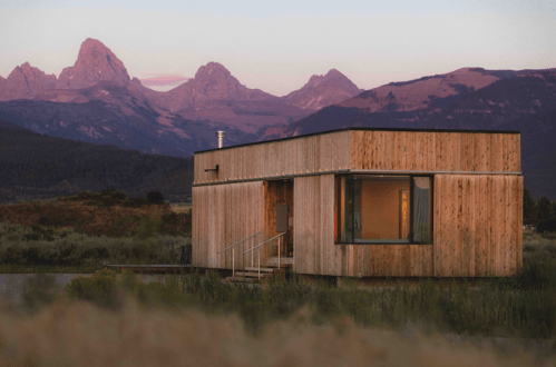 A modern wooden cabin surrounded by grass, with mountains in the background during sunset.