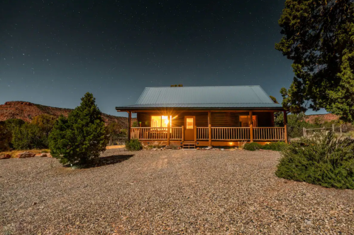 A cozy cabin with a lit window, surrounded by gravel and trees, under a starry night sky.