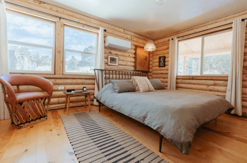 Cozy log cabin bedroom with a bed, wooden chair, and large windows letting in natural light.