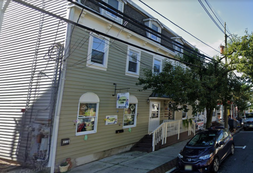 A two-story green building with white trim, featuring large windows and a staircase, located on a street with parked cars.