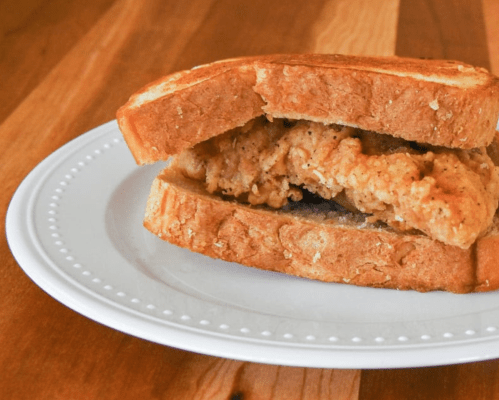 A crispy fried chicken sandwich on toasted bread, served on a white plate with a wooden background.