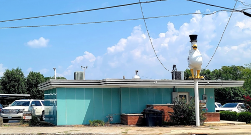 A turquoise building with a large white duck statue wearing a top hat on the roof, surrounded by parked cars.