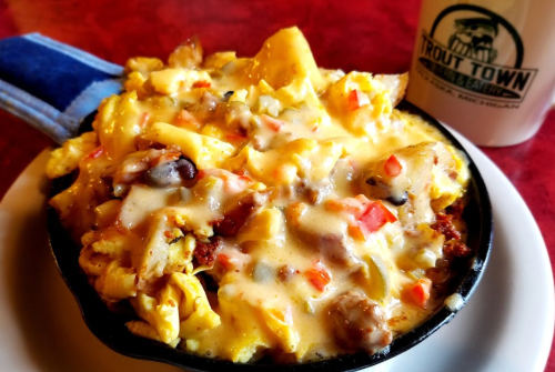 A sizzling skillet filled with cheesy scrambled eggs, peppers, and sausage, served with a coffee mug in the background.