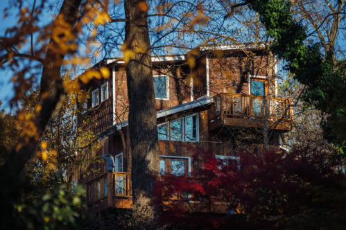 A multi-level wooden house nestled among trees, surrounded by autumn foliage and bright blue sky.