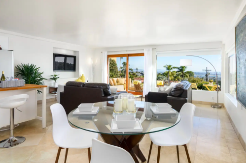 Modern living space with a glass dining table, white chairs, and a view of palm trees and mountains through large windows.