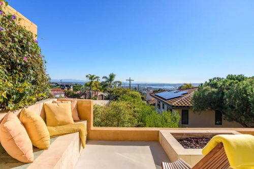 A sunny outdoor seating area with cushions, overlooking a scenic view of trees and distant hills under a clear blue sky.