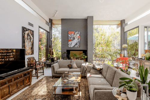 A modern living room with large windows, a gray sofa, artwork on the walls, and plants, creating a bright, inviting space.