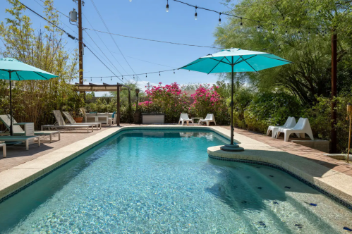 A serene pool area with turquoise umbrellas, lounge chairs, and vibrant flowers under a clear blue sky.