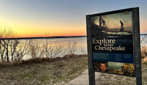 Sign reading "Explore your Chesapeake" by a calm waterway at sunset, with trees and grass in the foreground.