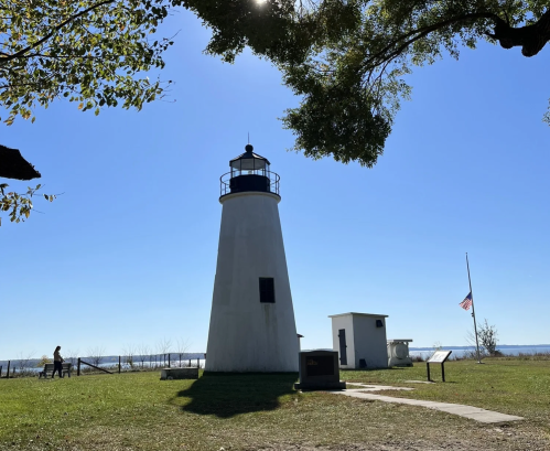 A white lighthouse stands against a clear blue sky, surrounded by trees and a grassy area, with a flag at half-mast.