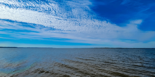 A serene view of calm water under a blue sky with scattered clouds, stretching to the horizon.