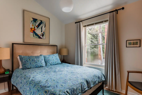 Cozy bedroom with a blue patterned bedspread, framed artwork, and natural light from a window.