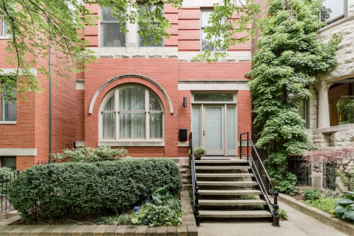 A modern brick building entrance with large windows, surrounded by greenery and steps leading to the door.