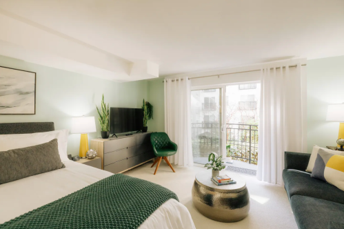 Cozy bedroom with a green accent, featuring a bed, TV, sofa, and large windows with natural light.