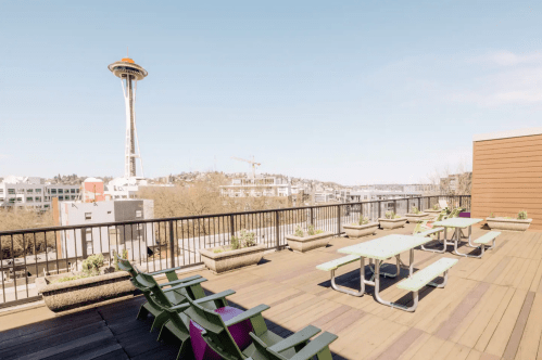 A rooftop terrace with picnic tables and chairs, featuring a view of the Space Needle and city skyline.