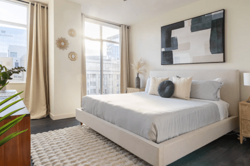 Bright, modern bedroom with a large bed, decorative pillows, and natural light from large windows. Stylish decor and plants.