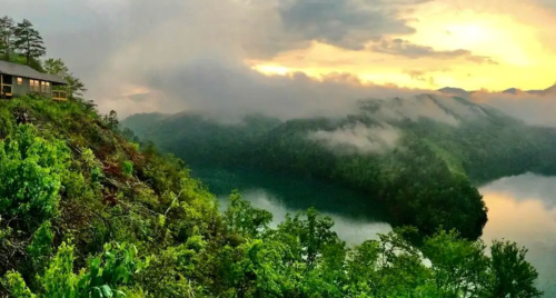 A scenic view of a misty lake surrounded by lush green hills and a cabin at sunset.