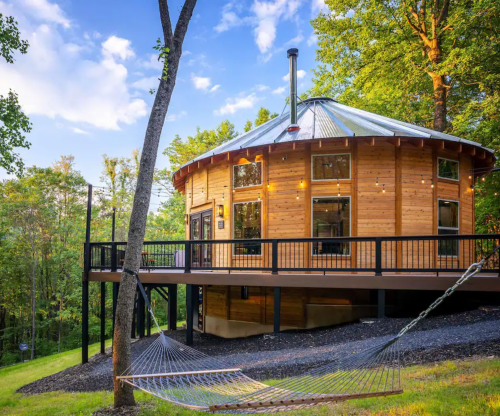 A round wooden cabin with a metal roof, surrounded by trees, features a deck and a hammock in a serene outdoor setting.