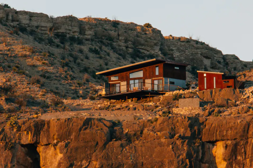 A modern wooden house perched on a rocky cliff, surrounded by rugged terrain and bathed in warm sunlight.