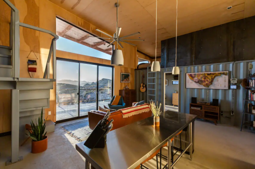 Modern living space with large windows, a spiral staircase, and a mix of wood and metal decor. Mountain view outside.