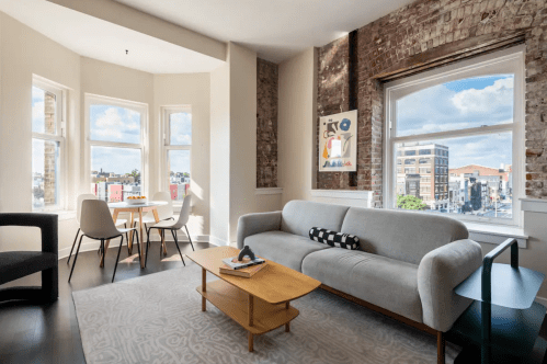 Bright living room with a gray sofa, wooden coffee table, and dining area by large windows showcasing a city view.