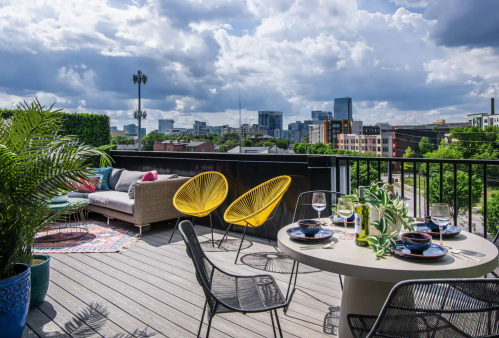 A stylish rooftop patio with yellow chairs, a table set for dining, and a view of a city skyline under a cloudy sky.