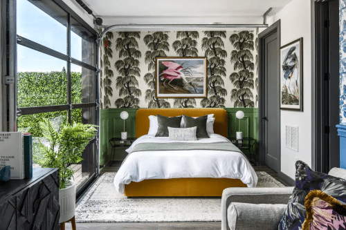 A stylish bedroom featuring a yellow bed, tropical wallpaper, and large windows with greenery outside.
