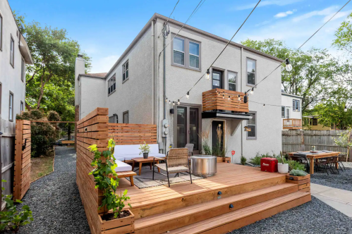A modern backyard with a wooden deck, seating area, and string lights, surrounded by greenery and gravel pathways.