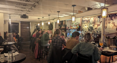 A cozy bar scene with patrons seated at tables and the bar, illuminated by warm pendant lights.