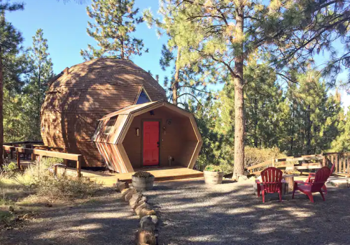 A unique wooden cabin with a round shape and an octagonal extension, surrounded by trees and outdoor seating.